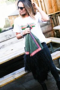 Claire Elizabeth standing holding green coat with black faux fur trim and wearing Taylor Morris sunglasses while holding her hair