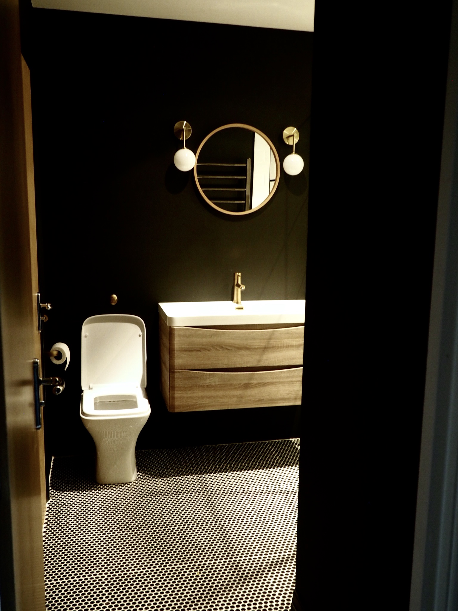 Bathroom entrance after makeover shower new white back to wall toilet and wide natural wood vanity unit sitting underneath two gold and white wall sconces and a round wood mirror