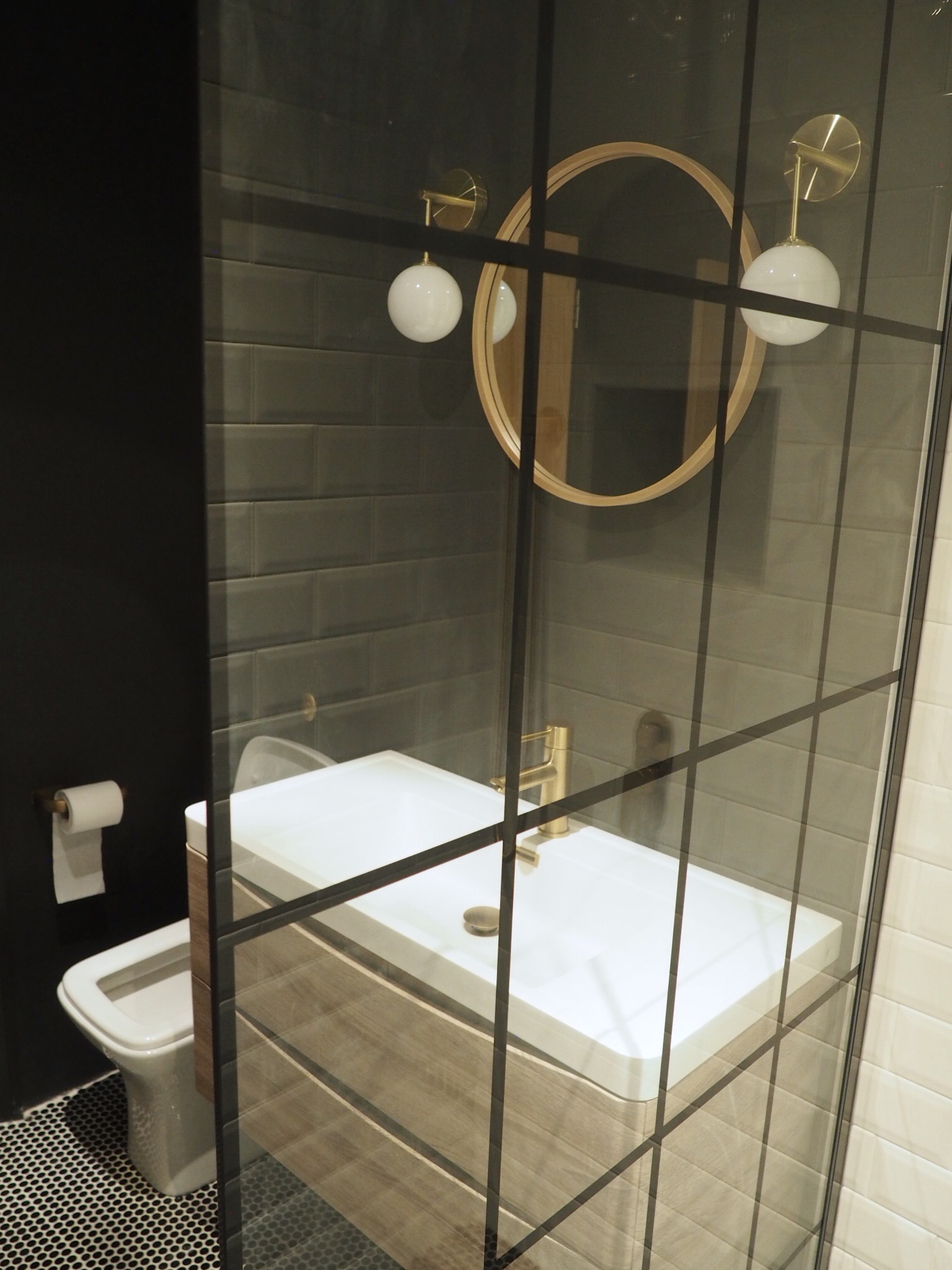 View from inside the shower showing the Crittal style shower screen and natural wood vanity unit with round wood Zara Home mirror and gold and white wall lights above