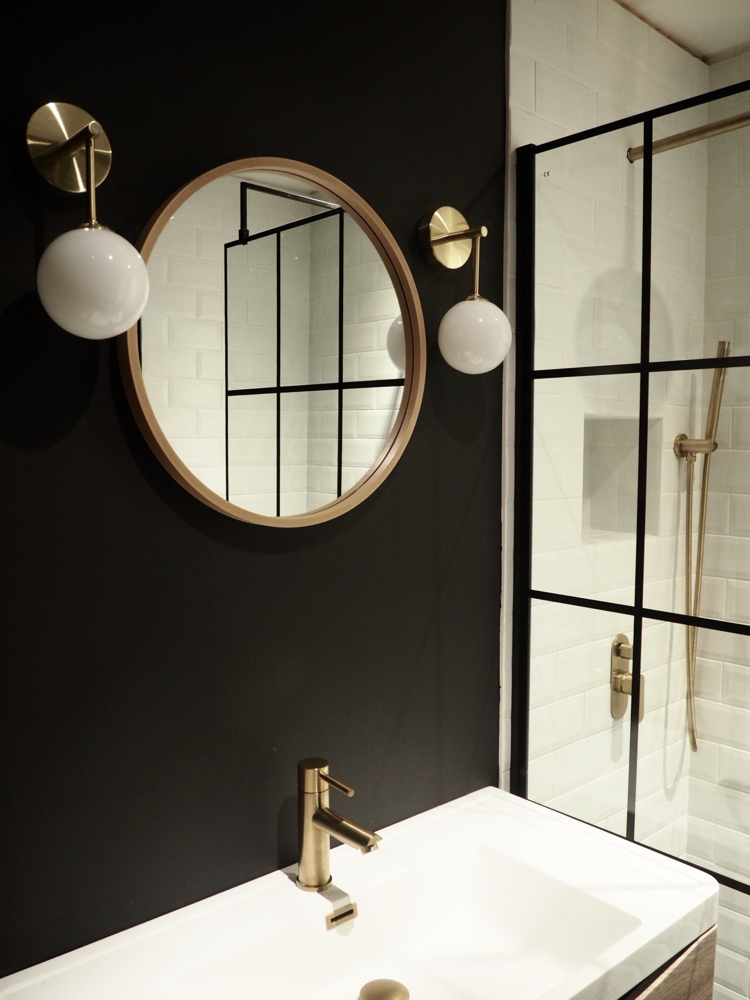 Natural wood vanity unit with round wood Zara Home mirror sitting above framed with two gold and white wall lights. Shower screen in Crittal style housing the brushed gold shower fittings and white subway tiles